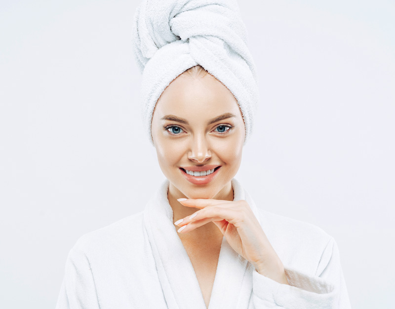 Fresh young woman wears bath towel and robe, touches chin gently, spends free time in spa