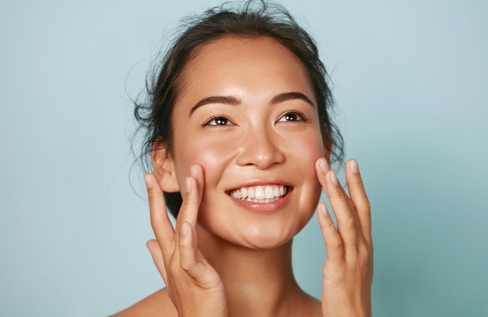 Smiling woman with glowing skin gently touching her face, representing facial treatments in Costa Mesa.