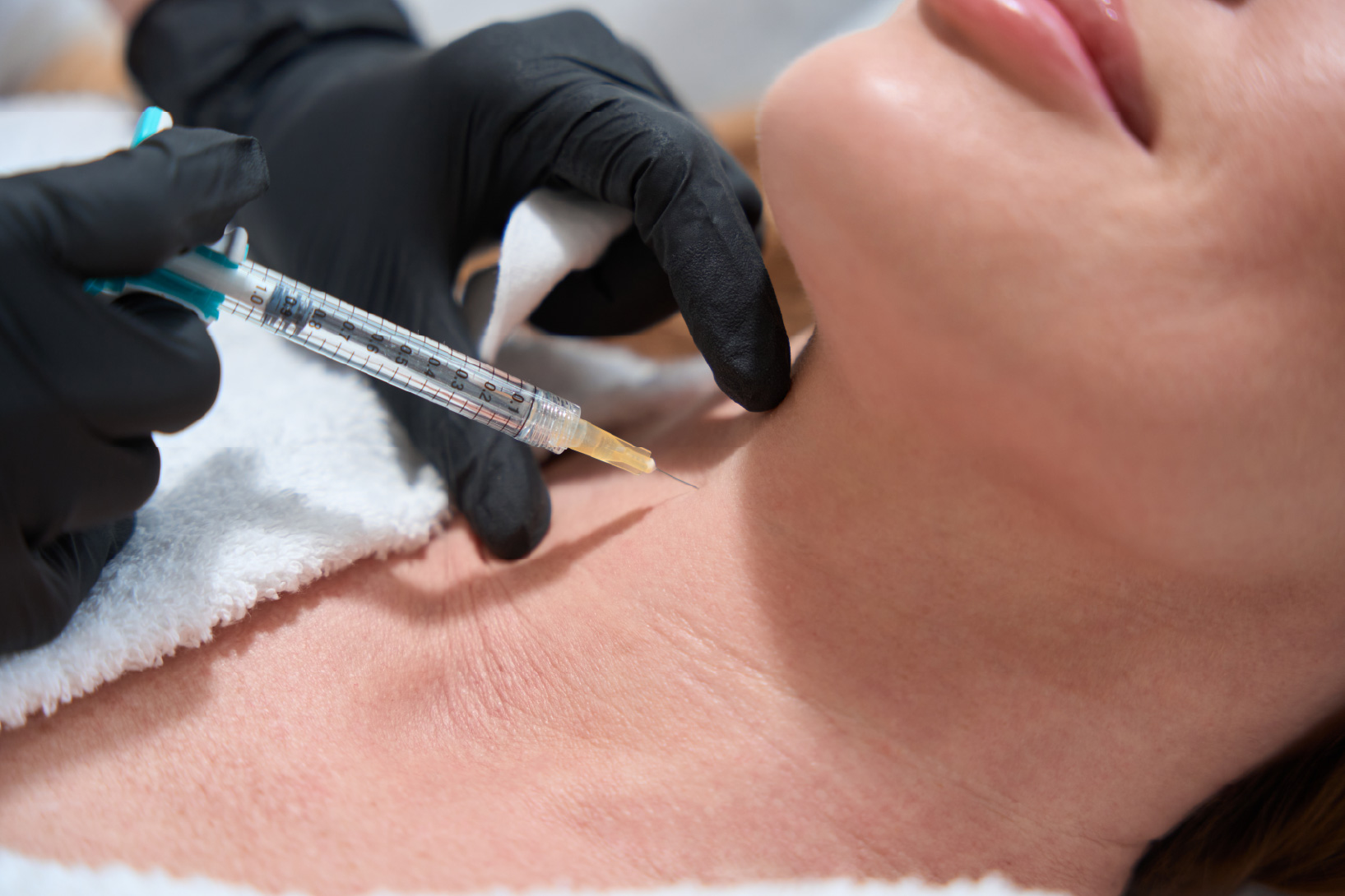 Gloved hands holding a syringe injecting the front of a woman’s neck while she lies with a towel under her chin.
