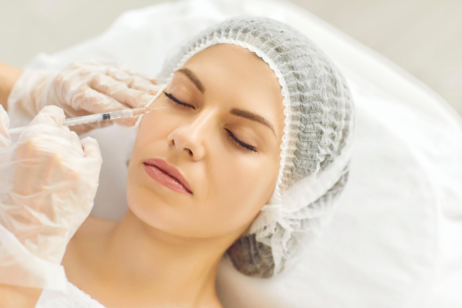 Woman lies with eyes closed while a gloved practitioner holds a syringe near her under-eye area.