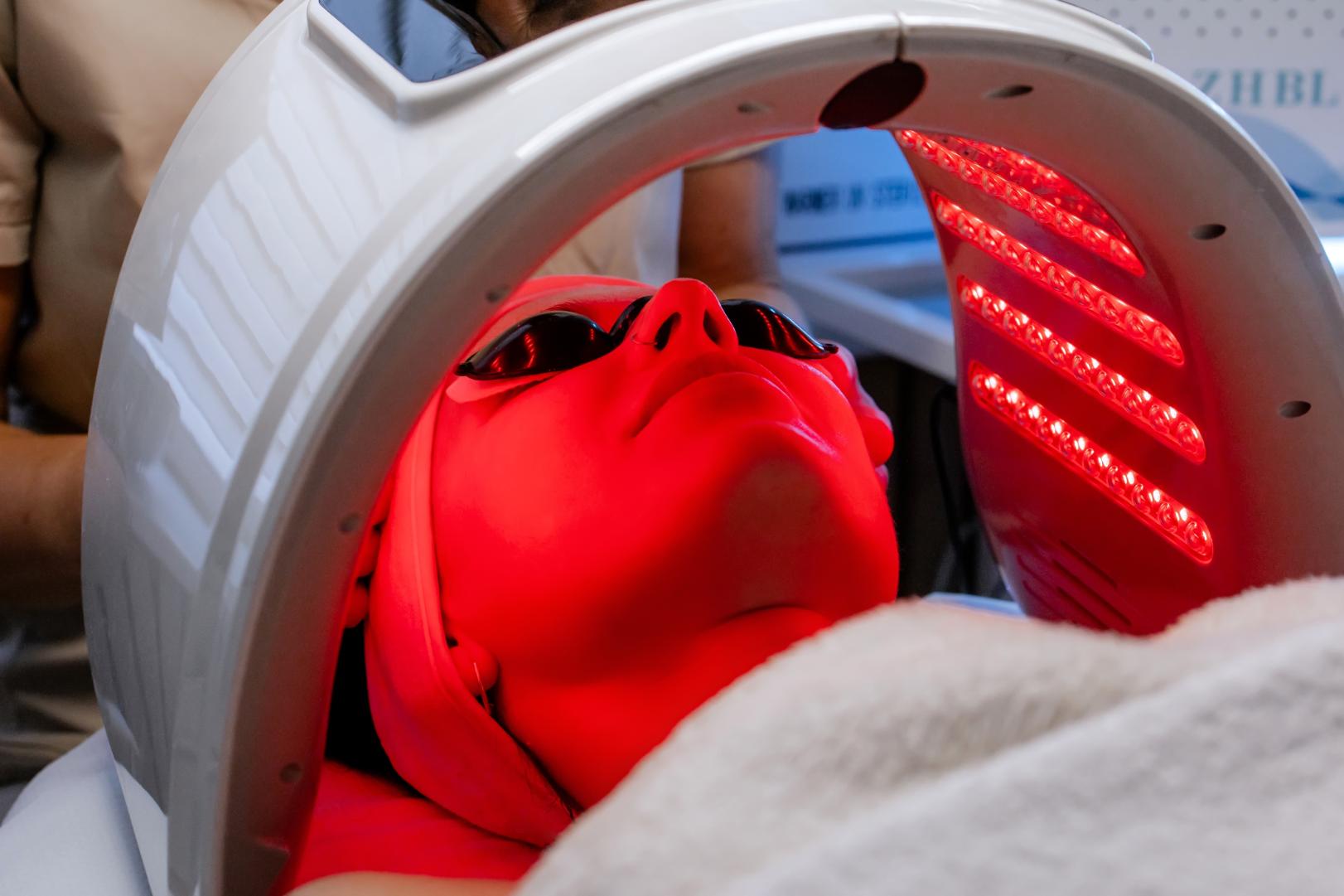 Woman wearing dark eye covers lies beneath a curved device with rows of red lights directed at her face.