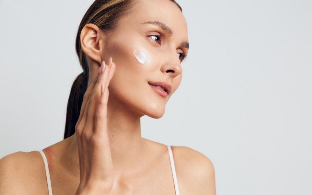 Woman applies a small amount of white skincare cream to her cheek against a light background.