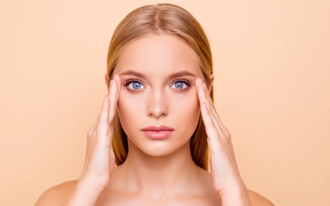 Woman facing forward with both hands placed near her temples against a peach background.